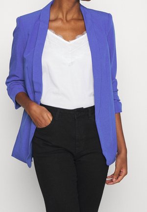 Une personne portant un blazer violet ajusté sur un haut blanc et un pantalon noir se tient avec une main dans la poche, mettant en valeur une tenue stylée.