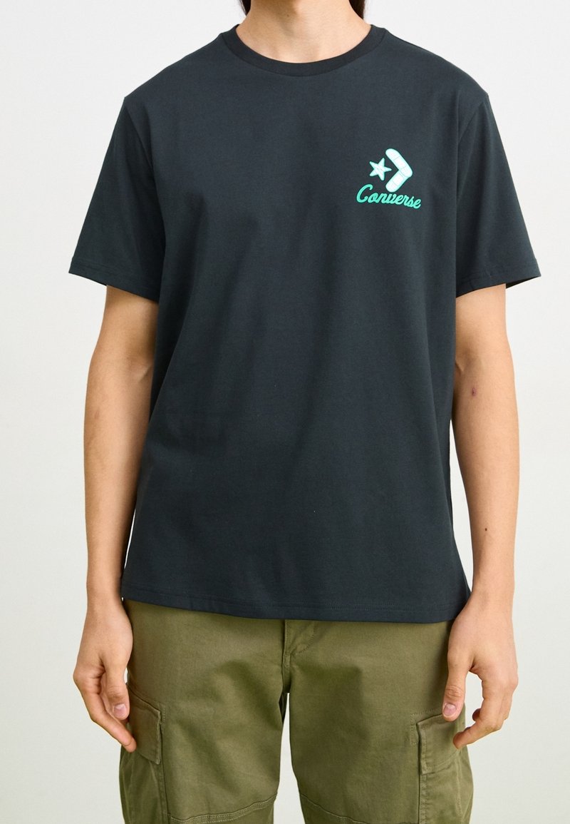 T-shirt en coton noir avec un graphisme avion et étoile en bleu sarcelle clair, col rond et manches courtes. Porté avec un pantalon cargo olive.
