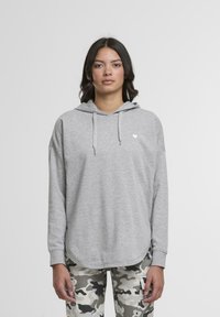 Szary hoodie z kapturem na sznurek, z małym białym logo serca na lewej piersi. Model zestawił go z spodniami w kamuflażu.