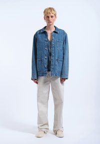 Giacca di jeans azzurra chiara con due tasche frontali, chiusura a bottoni e vestibilità rilassata, abbinata a pantaloni beige e sneakers beige.