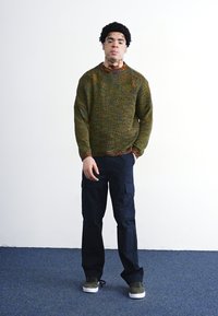 Sweter dzianinowy w ziemistych odcieniach z detalami faktury, w połączeniu z ciemnymi spodniami cargo. Zielone trampki dopełniają wygląd. Model stoi na jednolitym tle.