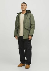 Zelena izolirana jakna s kapuco, dvema sprednjima žepoma, teksturirano blago in črnimi detajli. Nosi se z bež srajco in cargo hlačami.