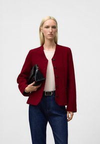 Czerwona kurtka z mieszanki wełny z okrągłym dekoltem, z przodu na guziki oraz długimi rękawami; zestawiona z beżową bluzką i ciemnoniebieskimi dżinsami.