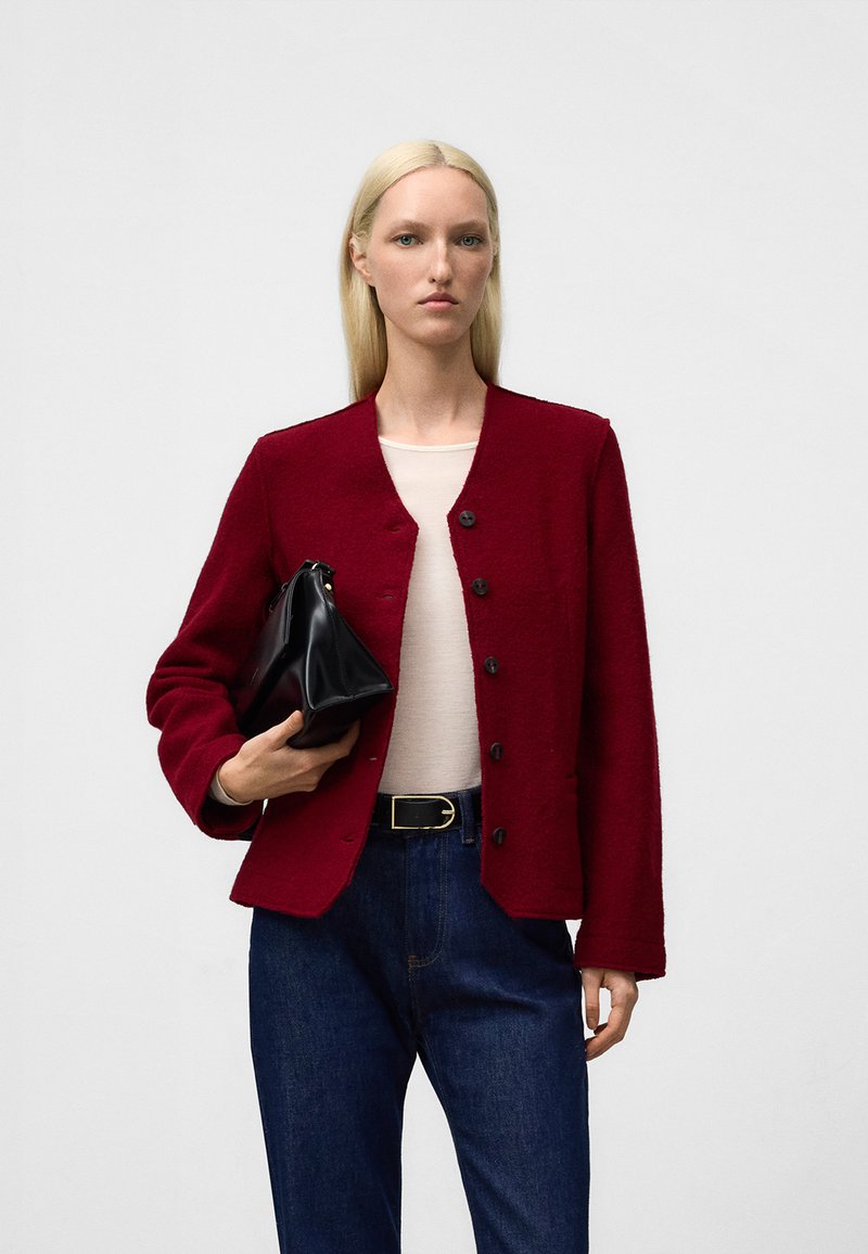 Czerwona kurtka z mieszanki wełny z okrągłym dekoltem, z przodu na guziki oraz długimi rękawami; zestawiona z beżową bluzką i ciemnoniebieskimi dżinsami.