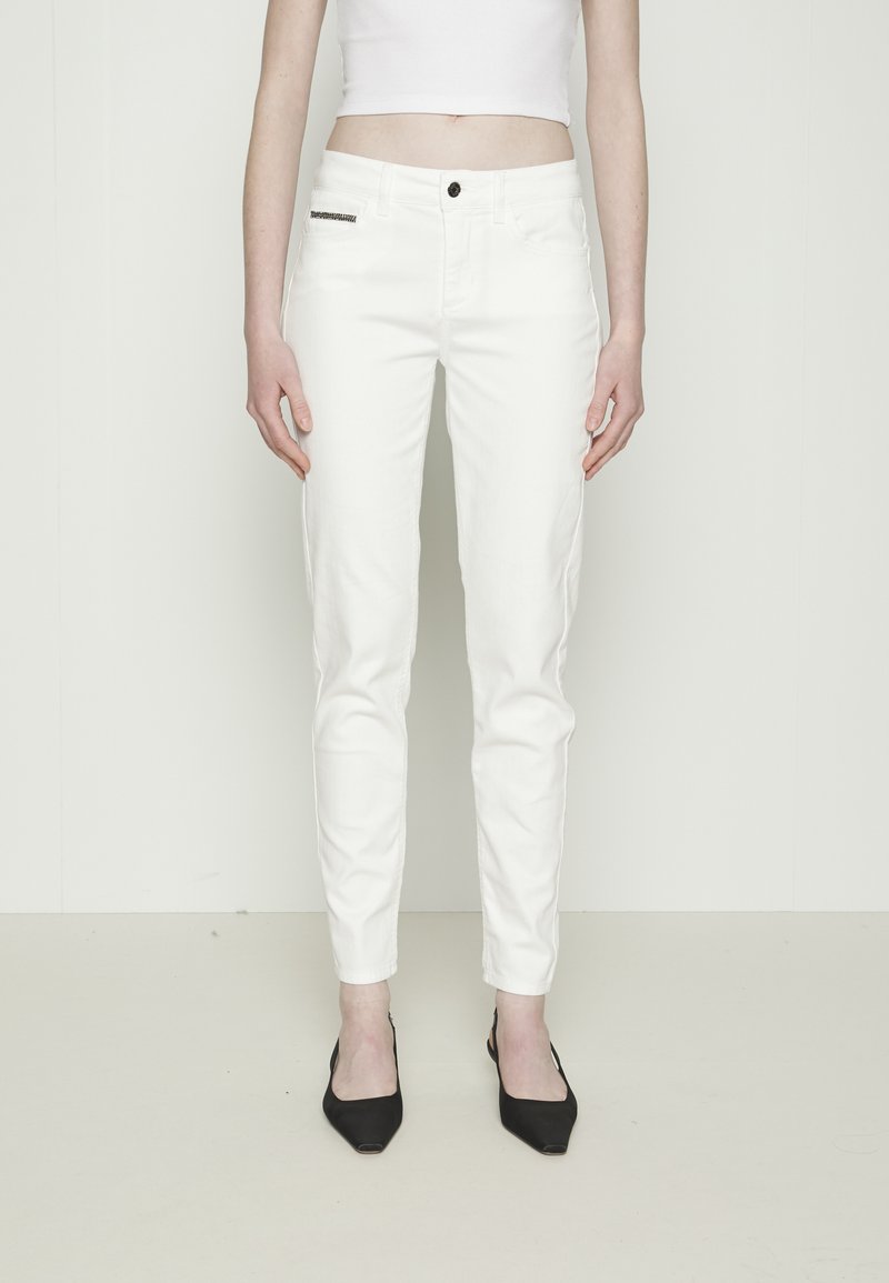 Femme portant un pantalon blanc slim, un top court blanc et des talons noirs pointus, debout devant un fond clair uni.