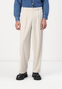 Pantaloni beige a gamba larga realizzati in tessuto testurizzato, con chiusura a bottone e tasche laterali, abbinati a scarpe con suola spessa nera.