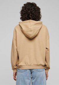 Sudadera beige con una textura suave, capucha grande, hombros caídos y puños acanalados. Combinada con unos vaqueros de denim de corte holgado en azul claro.