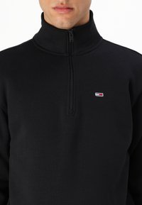 Zwarte zip-up hoodie met een hoge kraag, gemaakt van zacht materiaal. Voorzien van een klein geborduurd logo in het wit, rood en blauw op de borst.
