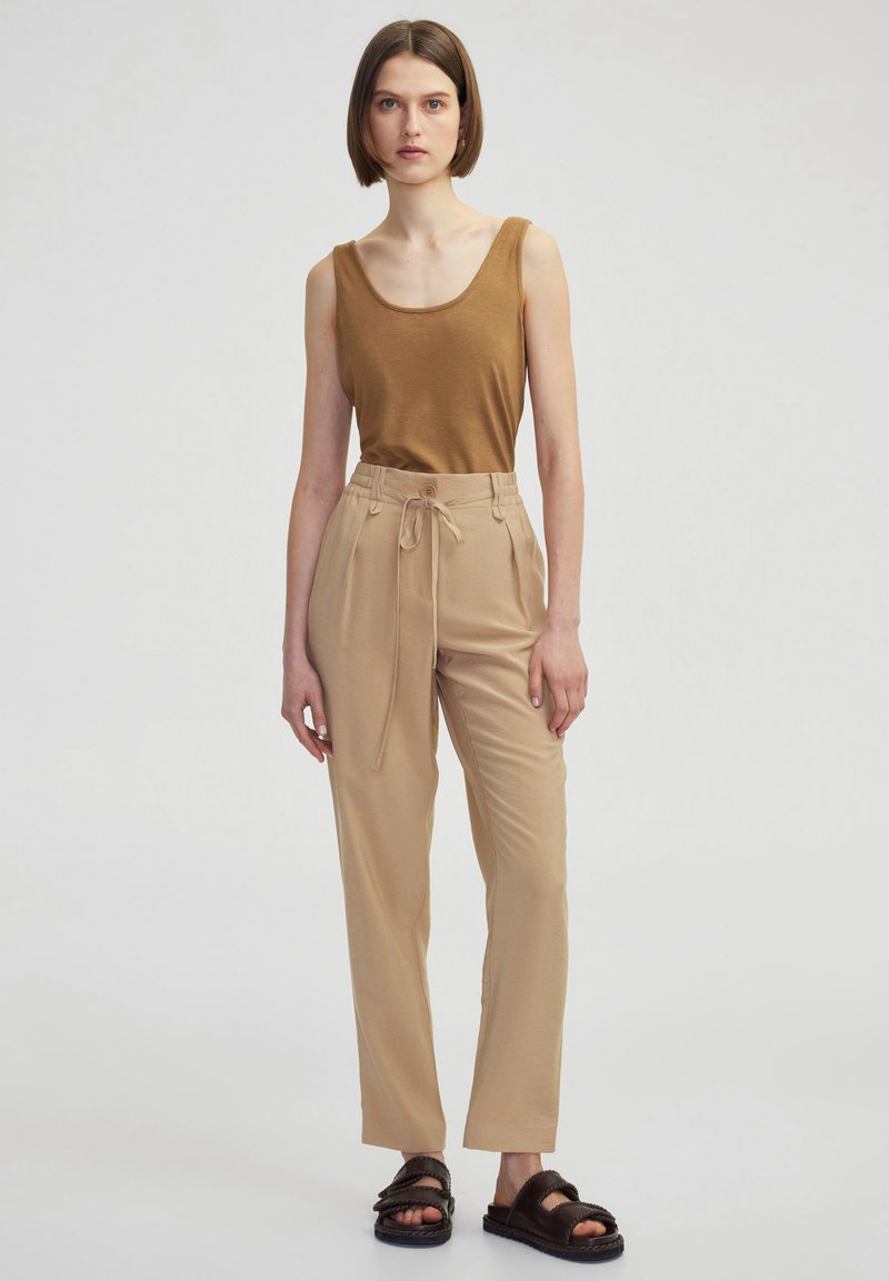Tan mouwloos topje, beige hooggeremde broek met strikceintuur, en donkere sandalen met gevlochten detail. Eenvoudige achtergrond.