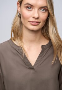 Brun bluse med V-hals og glat tekstur, der har en afslappet pasform og minimale syningsdetaljer. Modellen har bølget blont hår.