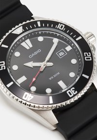 Montre analogique noire avec un boîtier rond en acier inoxydable, un bracelet en caoutchouc, des index des heures blancs, une aiguille des secondes rouge et une résistance à l'eau allant jusqu'à 200 mètres.