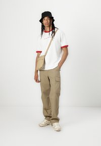 White t-shirt with red trim, beige cargo pants, and cream sneakers. Black bucket hat and beige crossbody bag complete the outfit.