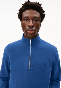 Polaire bleue à demi-fermeture éclair avec un col montant, dotée d'une fermeture éclair argentée et d'un petit logo brodé sur le côté gauche de la poitrine. Texture lisse.