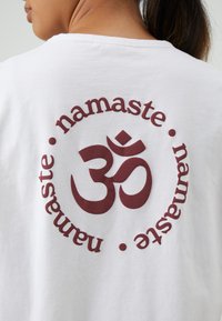 Bijela pamučna majica s velikim ljubičastim kružnim grafikom na kojoj se u središtu nalaze natpis "namaste" i simbol "ॐ". Mekana tekstura, ležeran dizajn.