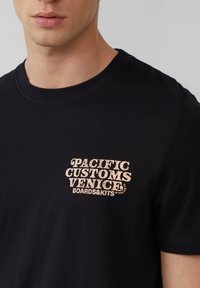 Zwarte katoenen t-shirt met een lichtbruine grafische logo op de borst dat "PACIFIC CUSTOMS VENICE BOARDS&KITS" leest. Klassieke pasvorm en korte mouwen.