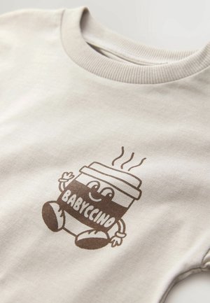 Tricou din bumbac bej cu un design imprimat de culoare maro, reprezentând o cană de cafea zâmbitoare, cu textul "BABYCINO" și linii de abur deasupra, guler texturat.