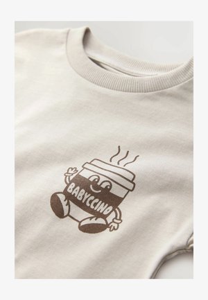 Bežs kokvilnas t-krekls ar brūnu apdrukātu dizainu, kurā attēlots smaidīgs kafijas krūze ar tekstu "BABYCINO" un tvaika līnijām virsū, ribota apkakle.