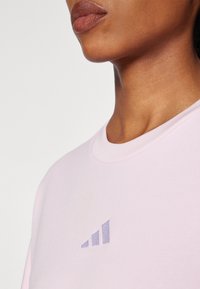 Lyse rosa sweatshirt med rund hals og ribbestrikket kant. Har et distinktivt lilla tre-stripes logo på brystet. Myk tekstur.