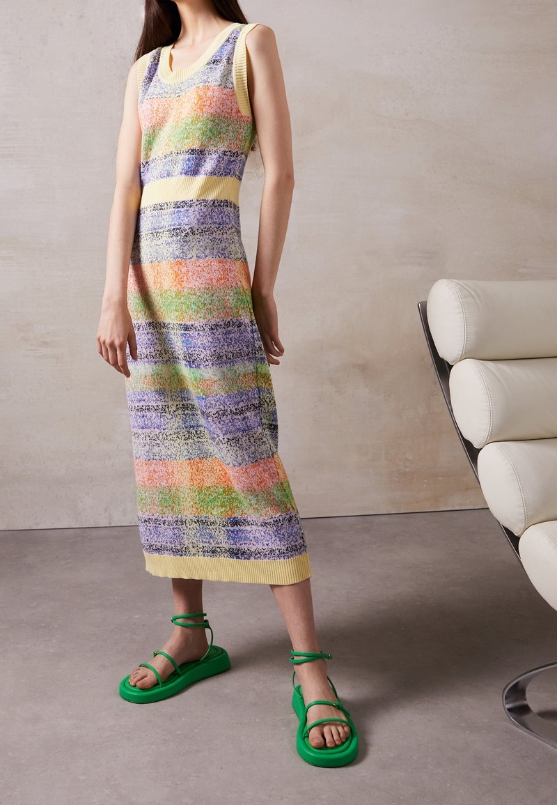 Femme portant une robe midi sans manches à rayures multicolores et des sandales vert vif, debout près d'une chaise moderne en cuir blanc.
