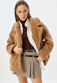 Giacca teddy in peluche marrone con ampi rever, sovrapposta a un maglione scuro a costine e una camicia bianca. Abbinata a una minigonna a quadri e una cintura nera.