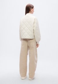 Μπουφάν σε κρεμ χρώμα με καπιτονέ λεπτομέρειες, κοντό μήκος, μανίκια μπαλούν και μοτίβο σε σχήμα ρόμβου. Συνδυασμένο με ανοιχτό μπεζ παντελόνι φαρδιάς γραμμής και λευκά αθλητικά παπούτσια.