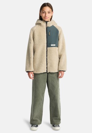 Giacca in pile beige con pannello turchese, chiusura con zip e tasche laterali, abbinata a pantaloni larghi verde oliva e scarpe da ginnastica.