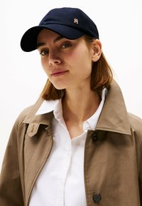 Marineblauw baseballpet met een gebogen rand en een gouden logo. Draagt bij een wit overhemd met kraag en een beige trenchcoat. Gladde texturen overal.