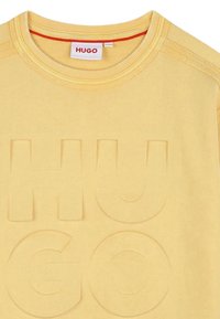 Sudadera amarilla con cuello redondo acanalado y logo "HUGO" en relieve. Hecha de algodón suave, presenta una textura lisa y costuras sutiles.