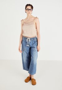 Top tricotat cu dungi în nuanțe de bej și alb, asortat cu pantaloni largi din denim albastru, care au talie elastică și șnururi. Clogs din suede maro.