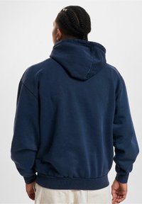 Granatowa bluza z kapturem o miękkiej fakturze, z dużym kapturem oraz prążkowanymi mankietami i dolnym wykończeniem. Widoczna od tyłu, z minimalnymi detalami projektu.