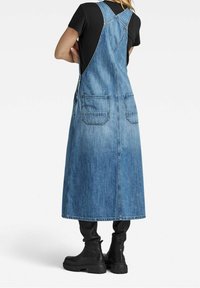 Denim overall dress in light blue, knee-length with two back pockets, adjustable straps, and a straight cut, worn over a black top and black boots.