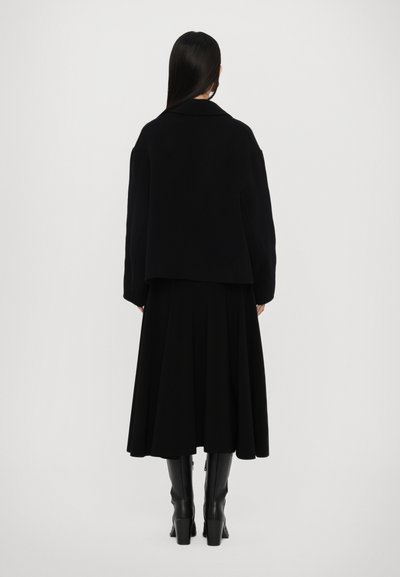 Schwarzer Wollmantel mit kastiger Silhouette und breitem Kragen, getragen über einem schwarzen Rock, kombiniert mit knielangen schwarzen Stiefeln. Glatte Textur.