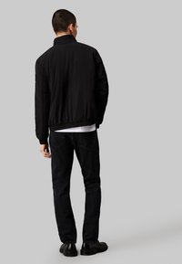 Man som bär en svart jacka, svarta jeans och svarta skor, står med ryggen mot en neutral grå bakgrund.