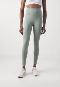 Leggings a vita alta e aderenti in morbido tessuto verde oliva. Presentano una superficie liscia e cuciture minime, abbinate a scarpe sportive bianche.