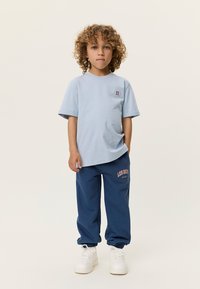 Ανοιχτό μπλε κοντομάνικο μπλουζάκι με ένα μικρό κόκκινο σχέδιο, σεταρισμένο με σκούρο μπλε παντελόνι φόρμας που έχει ροζ κείμενο, ελαστικές μανσέτες και λευκά αθλητικά παπούτσια.