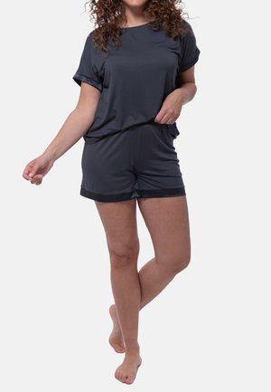 Vrouw staat blootsvoets, draagt een donkergrijze T-shirt met korte mouwen en bijpassende short tegen een effen lichte achtergrond.