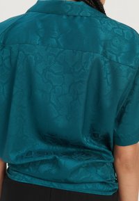 Türkisfarbendes Satin-Hemd mit kurzen Ärmeln und dezentem Schlangenmotiv, getragen von einer Person mit geflochtenem Haar, von hinten betrachtet.