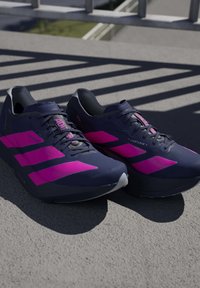 Paire de chaussures de course bleu marine avec des bandes rose vif posées sur une surface en béton gris avec des motifs d'ombre provenant d'une rambarde.