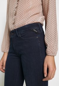 Dark blue jeans with a smooth texture and fitted design. Accompanied by a light, polka-dotted blouse with a subtle sheen and button details.