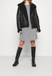 Musta nahkatakki, jossa on pörröinen kaulus ja hihansuut, yllä kuvioitu harmaa mekko. Mallilla on polveen asti ulottuvat mustat saappaat.