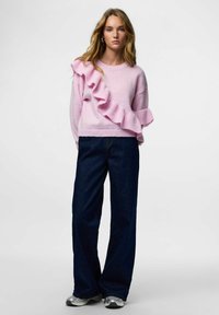 Jasnoróżowy dziergany sweter z dużym falbanowym akcentem po lewej stronie, w połączeniu z ciemnoniebieskimi szerokimi spodniami i srebrnymi trampkami.