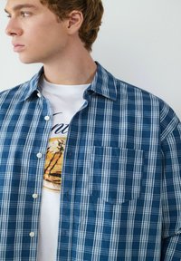 Camicia a quadri blu e bianca con taschino sul petto e morbido materiale in cotone, indossata sopra una maglietta bianca con grafica.