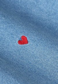 Tissu en denim bleu clair, avec un petit cœur brodé rouge. Le matériau présente une texture lisse avec des motifs de tissage visibles.