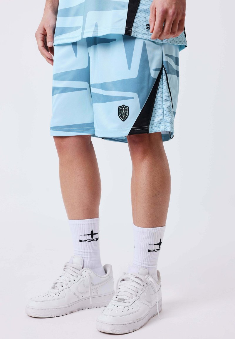 Homme portant un short bleu clair à motifs avec des fentes latérales, des chaussettes blanches de marque à tige courte et des baskets blanches Nike, debout sur un fond clair.