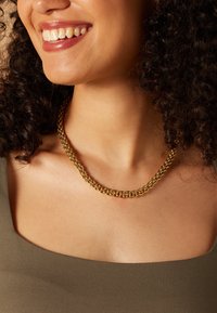 Gouden kettinghalsband met meerdere gevlochten strengen, die het licht reflecteren; gedragen op een blote nek met een ronde, olijfgroene top.