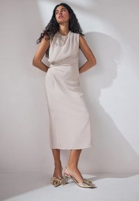 Robe sans manches beige avec un design drapé et un nœud sur le côté. Chaussures métalliques avec un bout pointu et un accent en nœud. Texture lisse.