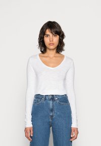 Balta ilgomis rankovėmis palaidinė su V formos iškirpte, lengvos tekstūros, derinama su aukštu liemeniu mėlynomis džinsomis. Paprastas, kasdienis dizainas su laisvu siluetu.