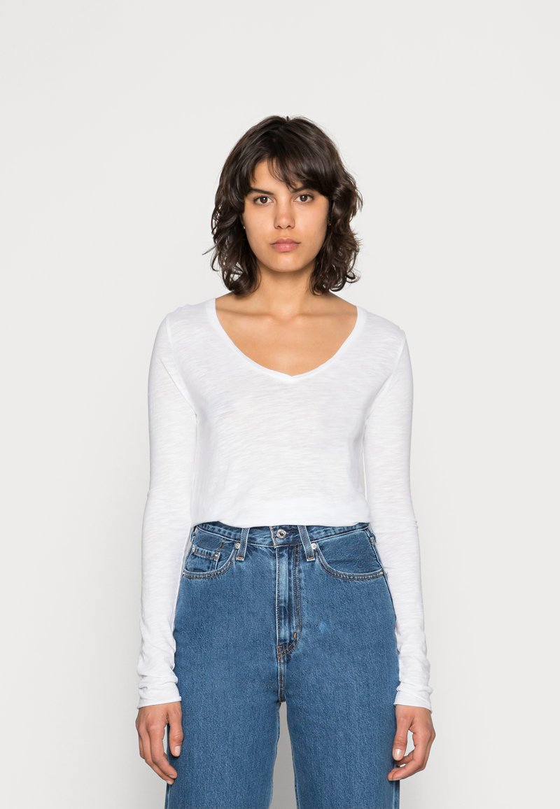 Balta ilgomis rankovėmis palaidinė su V formos iškirpte, lengvos tekstūros, derinama su aukštu liemeniu mėlynomis džinsomis. Paprastas, kasdienis dizainas su laisvu siluetu.