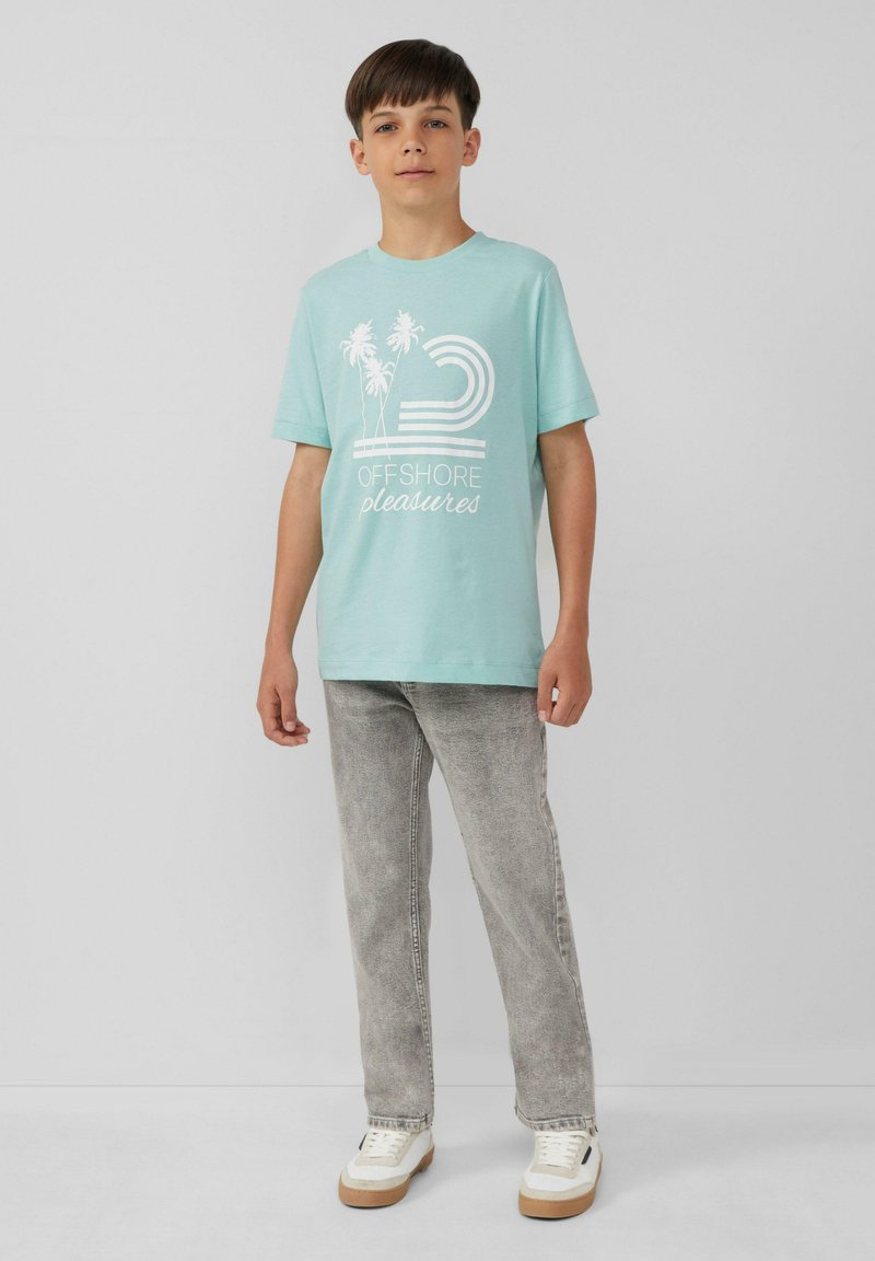 Hellblaues Baumwoll-T-Shirt mit weißem Grafikdesign, das Palmen und Text zeigt. Kombiniert mit grauen Denim-Jeans und weißen Turnschuhen.
