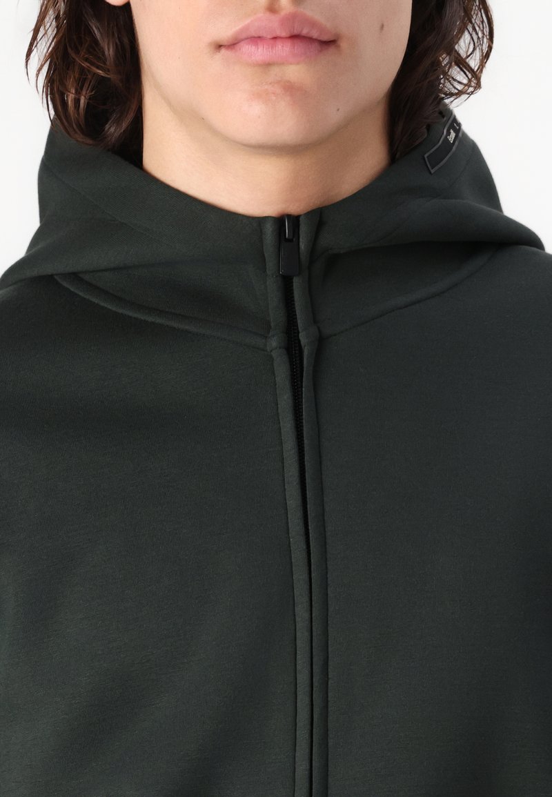 Młoda osoba z kręconymi włosami, ubrana w ciemnozieloną bluzę z kapturem z całkowicie zapiętym zamkiem błyskawicznym i podniesionym kapturem.